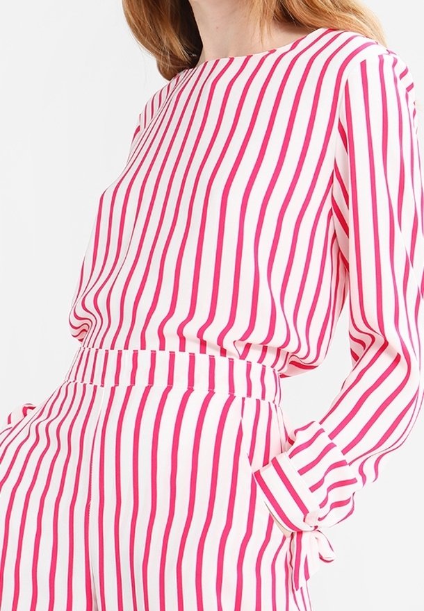 Vrouw in een roze en witte verticaal gestreepte blouse met lange mouwen en bijpassende hoge taillebroek, met haar hand in de zak.