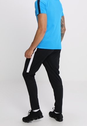 Mann trägt blaues kurzärmeliges Hemd und schwarze Sporthose mit weißem Streifen, steht mit teilweise gedrehtem Rücken, schwarze Sneakers auf weißem Boden.