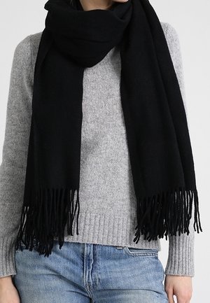 Personne portant un pull en maille gris et un jean bleu avec une longue écharpe noire à franges enveloppée autour du cou.