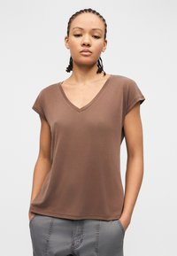 Νέος με πλεξούδες, φοράει καφέ μπλουζάκι με V και γκρι παντελόνι, στέκεται με τα χέρια στις τσέπες μπροστά σε απλό φόντο.