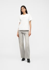 Γυναίκα με λευκό μπλουζάκι με κείμενο και ανοιχτό γκρι καμπάνα τζιν, στέκεται μπροστά σε απλό λευκό φόντο, φορώντας μαύρες μπότες με τακούνι.