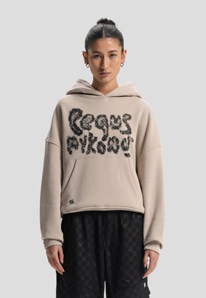 Young woman with dark hair in a bun wears a beige hoodie with black fuzzy text and black patterned loose pants against a plain gray background.