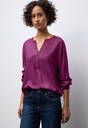 Magenta, langärmlige Bluse mit V-Ausschnitt, glatter Textur und gerafften Manschetten, kombiniert mit dunkelblauen Jeans mit Taschen-Details.