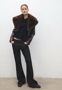 Chaqueta de cuero con collar de piel marrón sobre un suéter de punto azul marino, combinada con jeans negros de campana y calzado marrón oscuro, todo ello dispuesto sobre un fondo sencillo.