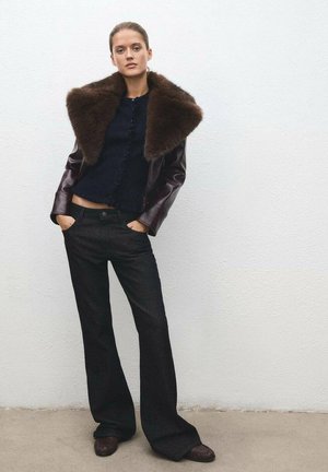 Veste en cuir avec col en fourrure marron sur un pull côtelé bleu marine, associée à un jean noir évasé et des chaussures marron foncé, le tout sur un fond uni.