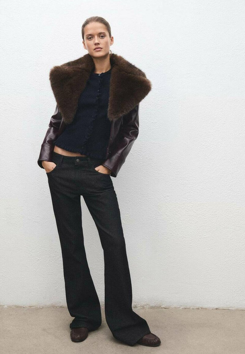Chaqueta de cuero con collar de piel marrón sobre un suéter de punto azul marino, combinada con jeans negros de campana y calzado marrón oscuro, todo ello dispuesto sobre un fondo sencillo.