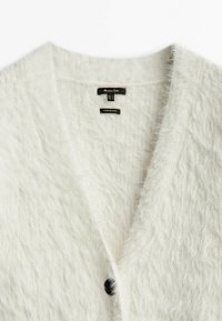 Cardigan bianco peloso con scollo a V e un singolo bottone nero. Etichette visibili che indicano marca e taglia. Texture morbida, design aperto.