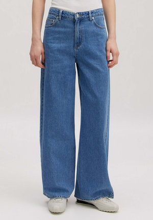 Personne portant un jean bleu taille haute ample, associé à des baskets blanches et un haut blanc, debout devant un fond uni.