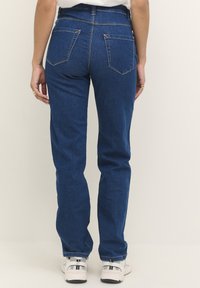 Jean en denim bleu taille haute avec une jambe droite, deux poches arrière et des coutures jaunes visibles. Portez avec des baskets blanches.