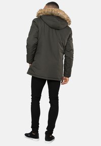 Hombre con parka verde oscuro con capucha forrada de piel, pantalones negros ajustados y zapatillas negras, de pie con la espalda hacia la cámara.