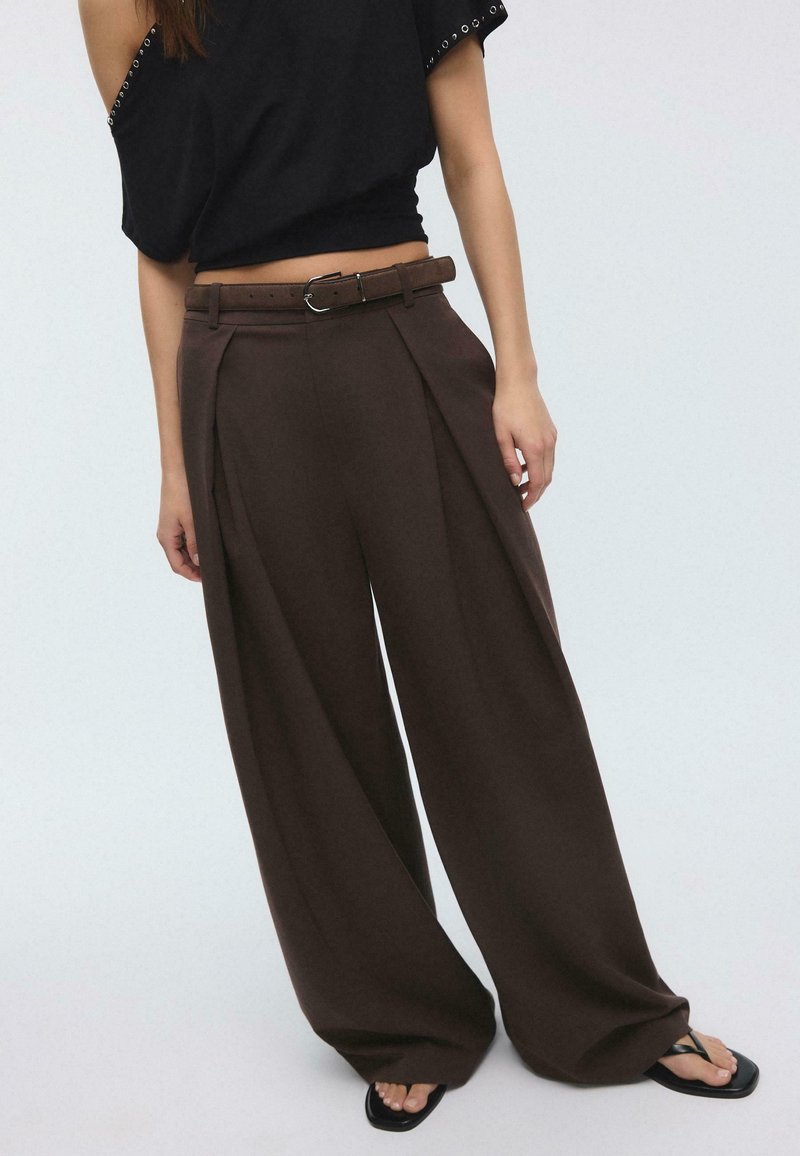 Woman wearing high-waisted wide-leg brown trousers with a matching belt, black cropped top, and black square-toe sandals.