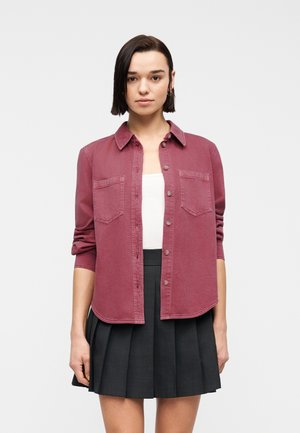 Frau trägt ein dunkelrotes Hemd mit Knöpfen über einem weißen Oberteil und einen plissierten schwarzen Rock, stehend vor einem schlichten hellen Hintergrund.