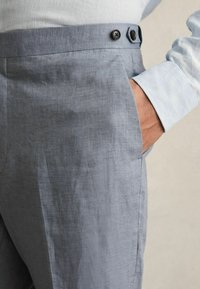 Pantalonii gri din in au o textură netedă, cu doi butoni negri la talie și o mână în buzunar pentru a oferi o idee de scară.