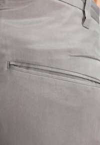 Gray fabric pant back featuring a concealed pocket with a smooth texture and simple stitching detail. Plain design without embellishments.