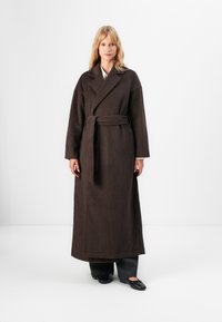 Brązowy, długi płaszcz wełniany z paskiem w talii, szerokimi klapami i długimi rękawami; ma fakturowaną powierzchnię i minimalistyczny design.