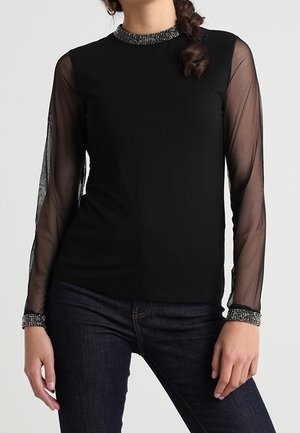 Vrouw draagt een zwarte top met doorschijnende lange mouwen en versierde manchetten en halslijn, gecombineerd met donkerblauwe spijkerbroek.