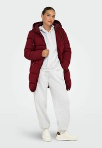 Abrigo acolchado burdeos con capucha, con textura acolchada y bolsillos con cremallera; combinado con una sudadera gris y pantalones de chándal, zapatillas blancas.