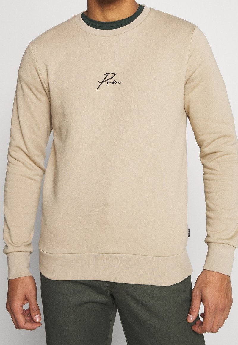 Beige collegepaita, joka on valmistettu pehmeästä kankaasta. Paitassa on pyöreä pääntie ja embroidered logo rinnassa. Elastiset hihansuut ja helma antavat rakenteen.