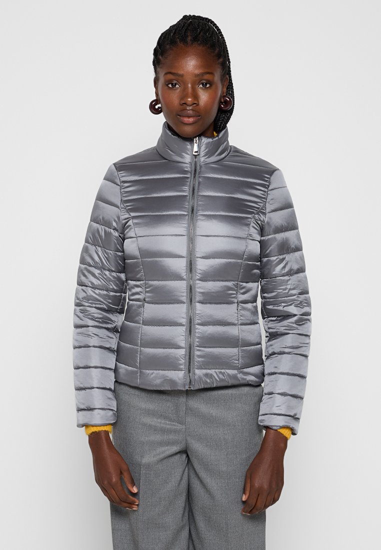 Frau in einer taillierten silbernen Pufferjacke, die zugezippt ist, mit grauen Hosen und sichtbaren gelben Pulloverärmel an den Handgelenken, steht vor einem schlichten Hintergrund.