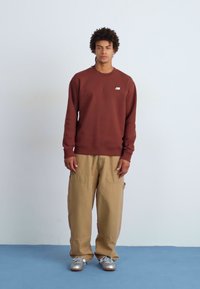 Brun genser med en liten hvit logo, kombinert med beige vide bukser og sølvfargede joggesko, stående mot en lyseblå bakgrunn.