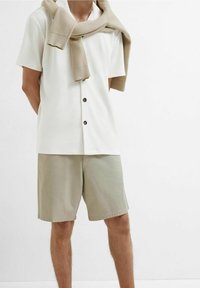 Person wearing a white short-sleeve buttoned shirt, beige shorts, and a beige sweater draped over shoulders against a white background.