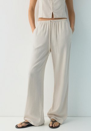 Žena oblečená v voľných béžových nohaviciach so sťahovacou šnúrkou, krátkom tielku bez rukávov a čiernych sandáloch, stojaca na jednoduchom svetlom pozadí.