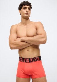 Mies, joka on ilman paitaa, kädet ristissä, päällä korallinpunaiset Calvin Klein -bokserit, joissa on musta vyötärönauha ja punainen logo valkoista taustaa vasten.