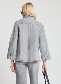 Veste en denim gris clair avec col, coupe oversize, coutures apparentes et poignets à boutons, portée avec un pantalon assorti gris clair.