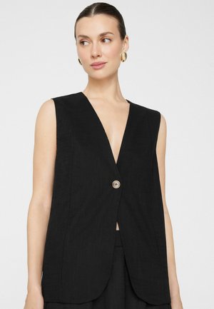 Femme portant un blazer noir sans manches avec un bouton, boucles d'oreilles en anneaux dorés, cheveux tirés en arrière, regardant légèrement vers sa gauche.