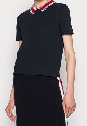 Osoba nosząca granatową bluzkę z krótkim rękawem z kołnierzykiem w czerwono-białe paski oraz pasującą granatową spódnicę z czerwono-białymi paskami po bokach.