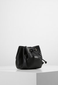 Černá kožená taška na stažení se skládanými boky, zlatými kovovými prvky a texturovaným povrchem, ležící na bílé platformě.