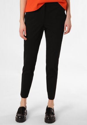 Personne portant un pantalon noir slim aux chevilles, des mocassins noirs épais et un haut orange sur un fond uni.