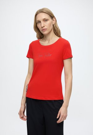 Blonde Frau mit Tattoos trägt ein rotes Kurzarmshirt mit dem Text "Tom Tailor" kombiniert mit schwarzen Hosen und steht vor einem schlichten Hintergrund.