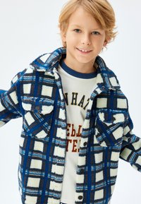 Chaqueta de forro polar a cuadros en azul, marrón y blanco con dos bolsillos en el pecho. Presenta un collar de camisa y una textura suave. Lleva una camisa beige debajo.