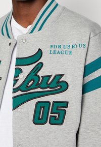 Giacca varsity grigia con dettagli in teal, caratterizzata da ricami con scritte e numeri, chiusura a bottoni a pressione e dettagli a righe sulle maniche.