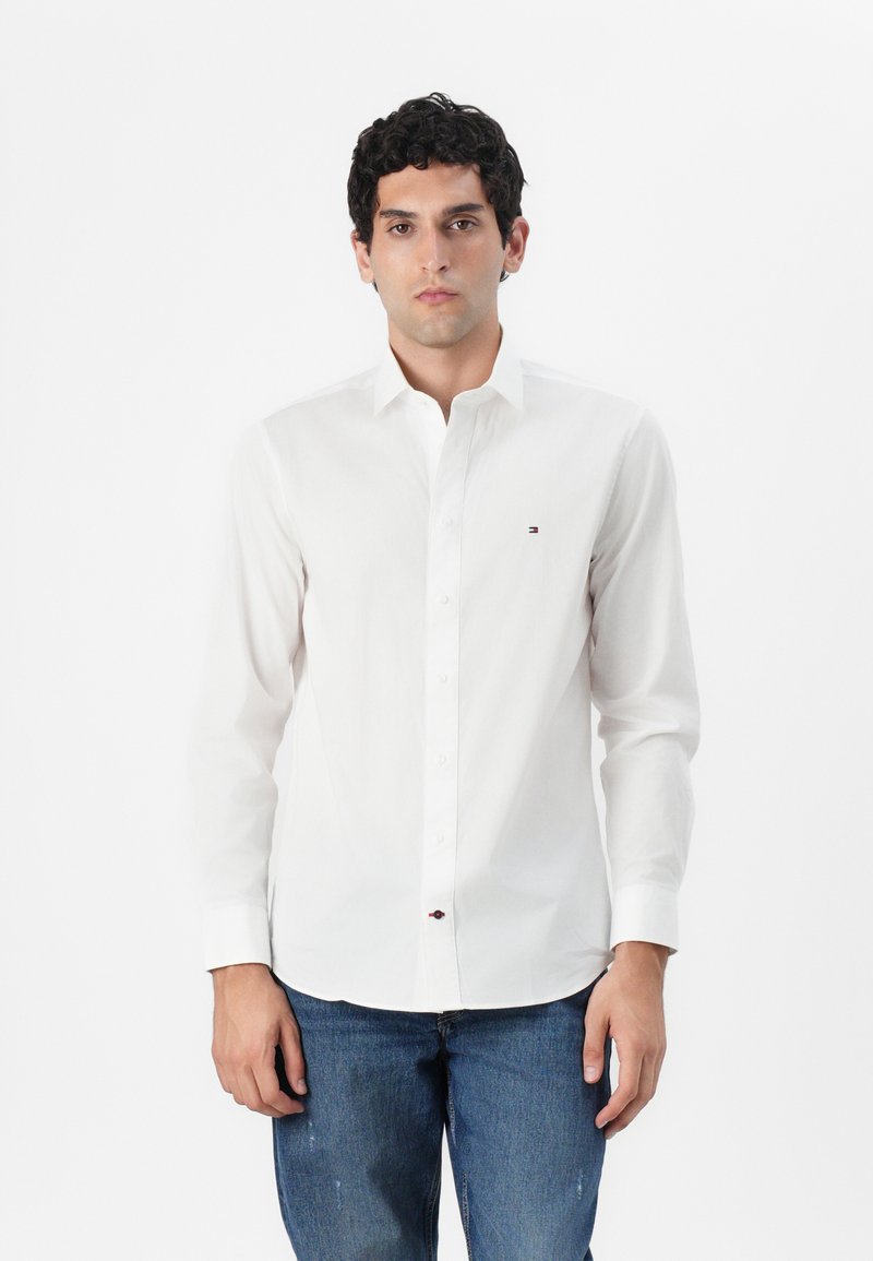 Chemise blanche à manches longues avec un col boutonné, ornée d'un petit logo sur la poitrine, assortie avec un jean en denim bleu.
