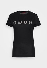 Camiseta negra de algodón con mangas cortas, que presenta letras metálicas invertidas que dicen "HUGO" en el pecho. Diseño simple.