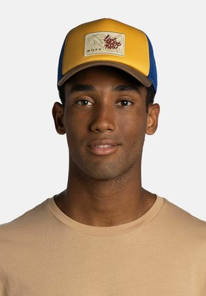 Junger Mann trägt beiges Hemd und gelb-blaue Kappe mit einem Berg-Patch mit der Aufschrift „live more now“ und „BUFF“ auf der Vorderseite.