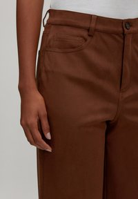 Braune Hosen aus glattem Stoff, gefertigt im klassischen Schnitt, mit Fronttaschen und Knopfverschluss. Die Textur wirkt weich und elegant.