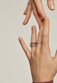 Anillos plateados y negros apilados en un dedo, con un delicado acento de gema en un anillo separado. Las manos tienen una textura de piel natural.