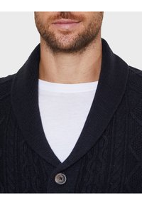 Navy knitted cardigan with a textured pattern, shawl collar, and a single button closure. Worn over a white crew-neck t-shirt.