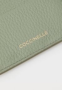 Green leather wallet with a textured finish, featuring gold "Coccinelle" branding and fine stitching detail along the edges.