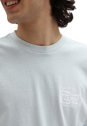 Jeune personne portant un T-shirt Vans bleu clair avec le logo "Off The Wall" sur la poitrine, souriant et regardant vers la gauche sur un fond blanc.