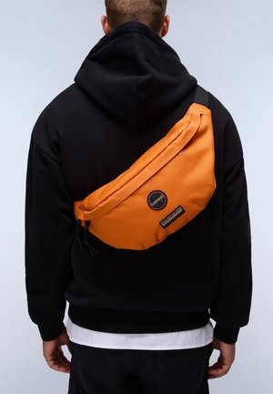 Orange sling bag with a curved shape, made of durable material. Features black accents, zippers, and a logo patch. Worn over a black hoodie.