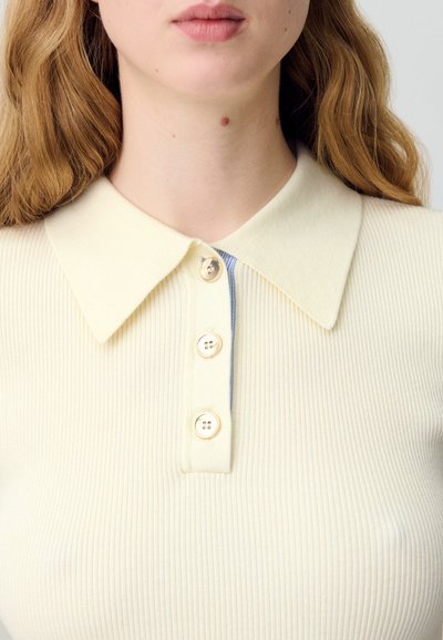 Polo de color amarillo pálido con canalé, que presenta un suave cuello y cuatro botones redondos de color claro en el frente, con un acento azul debajo.