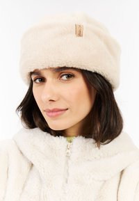 Woman wearing a cream-colored faux fur bucket hat and matching zip-up jacket, with a slight smile and shoulder-length dark hair.