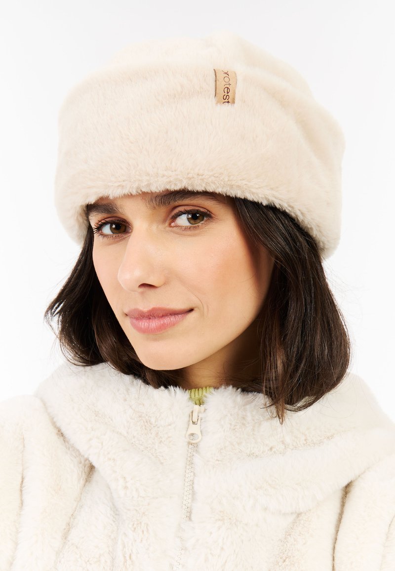 Woman wearing a cream-colored faux fur bucket hat and matching zip-up jacket, with a slight smile and shoulder-length dark hair.