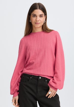 Femme aux cheveux bruns raides portant un haut rose texturé à manches longues et un jean noir taille haute, posant avec une main dans la poche.