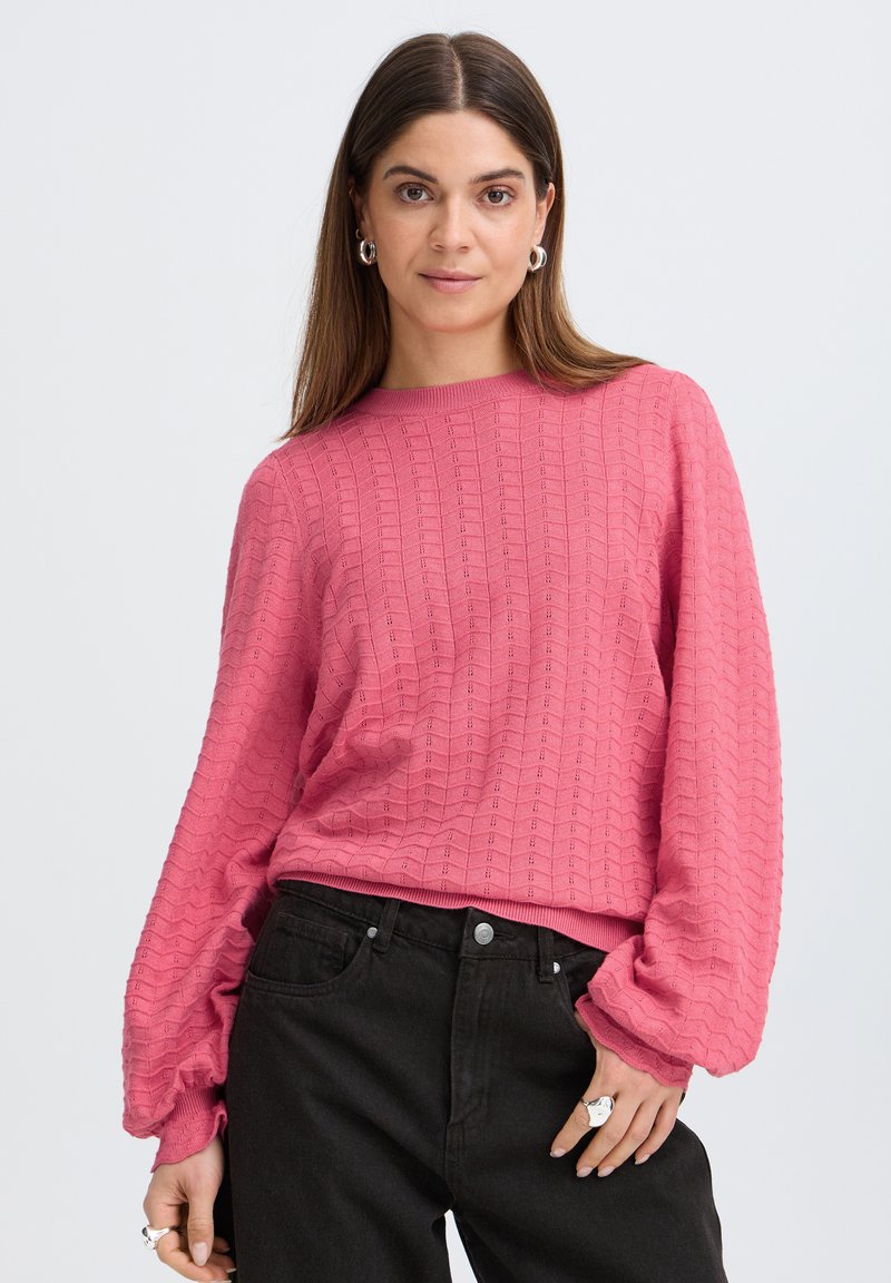 Femme aux cheveux bruns raides portant un haut rose texturé à manches longues et un jean noir taille haute, posant avec une main dans la poche.