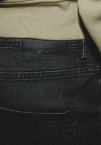 Jeans in denim nero con una etichetta in pelle che presenta il nome del marchio "diverse" sulla vita. Il tessuto ha una texture liscia.